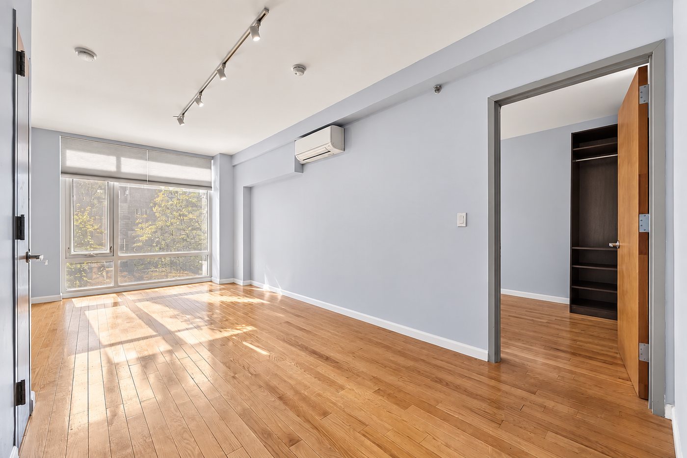 A sunlit, newly renovated bedroom with oak floors, pale blue walls, and a wall of windows onto trees.